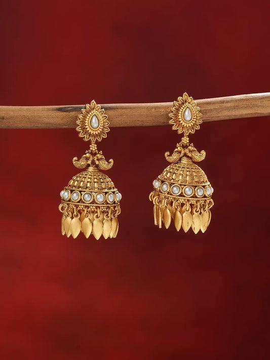 A pair of ornate gold-toned Indian jhumka earrings with white pearl-like accents, hanging from a wooden branch against a deep red background.