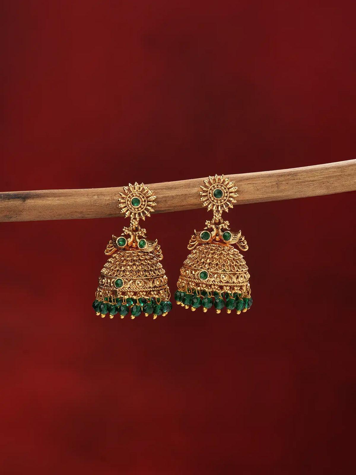 A pair of traditional Indian gold jhumka earrings with green stone accents and dangling green bead tassels, hanging from a wooden branch against a dark red background.