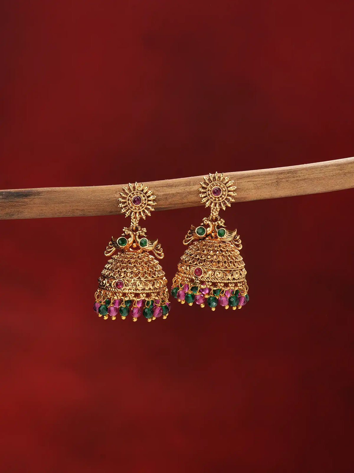 A pair of ornate gold-toned jhumka earrings with green and pink gemstone accents, hanging from a wooden branch against a dark red background.