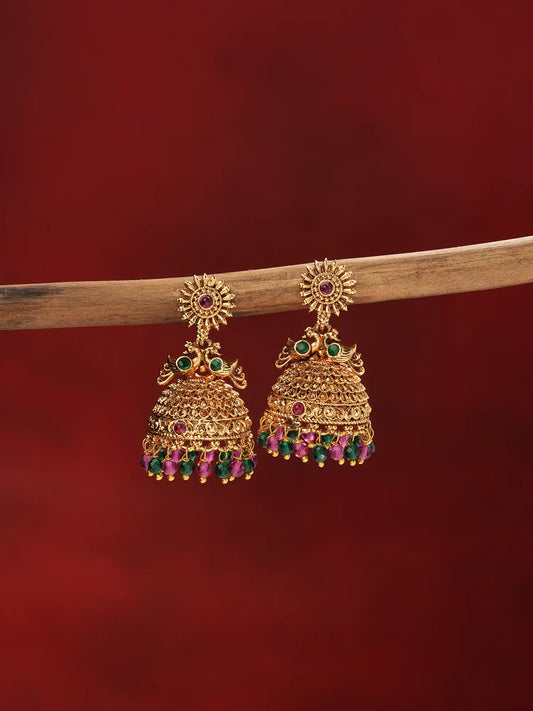 A pair of ornate gold-toned jhumka earrings with green and pink gemstone accents, hanging from a wooden branch against a dark red background.