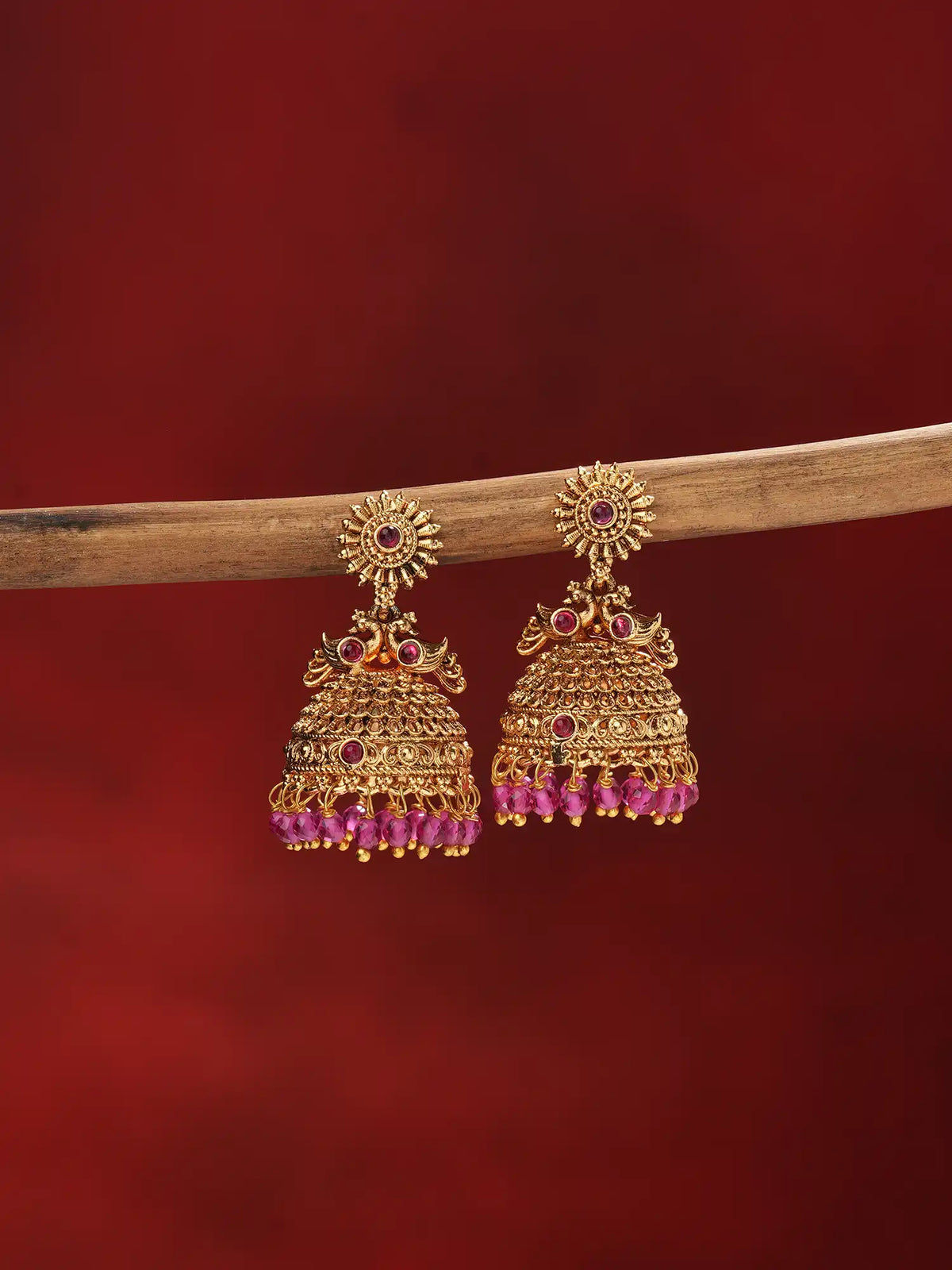 A pair of ornate gold-toned Indian jhumka earrings with red gemstone accents and dangling pink beads, hanging from a wooden branch against a dark red background.
