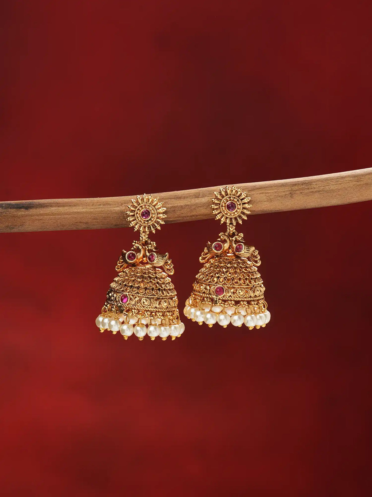 A pair of ornate gold-toned jhumka earrings with red stone accents and dangling pearl-like beads, hanging from a wooden branch against a deep red background.