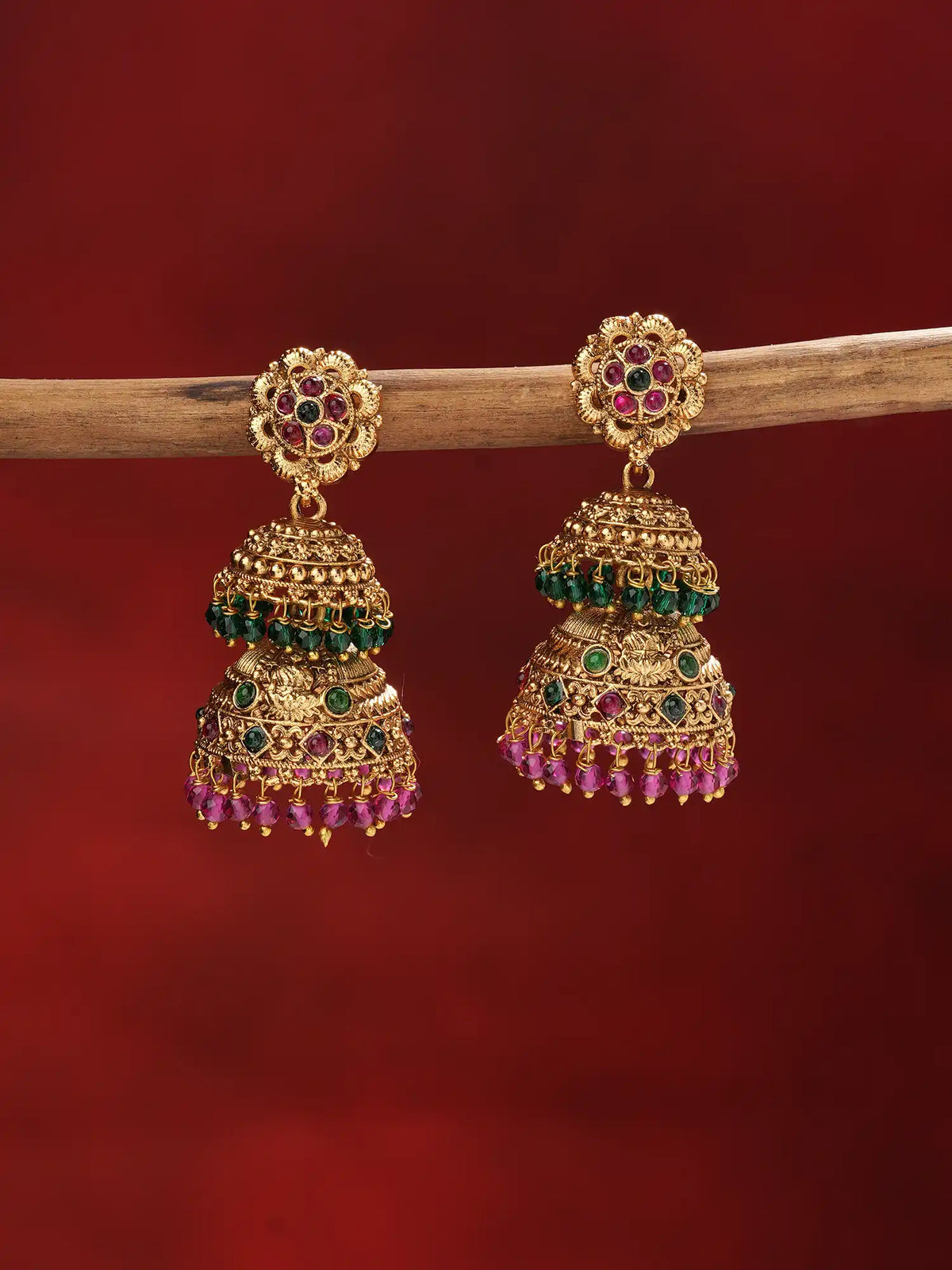 A pair of ornate gold-toned Indian jhumka earrings embellished with green and pink stones, hanging from a wooden branch against a dark red background.