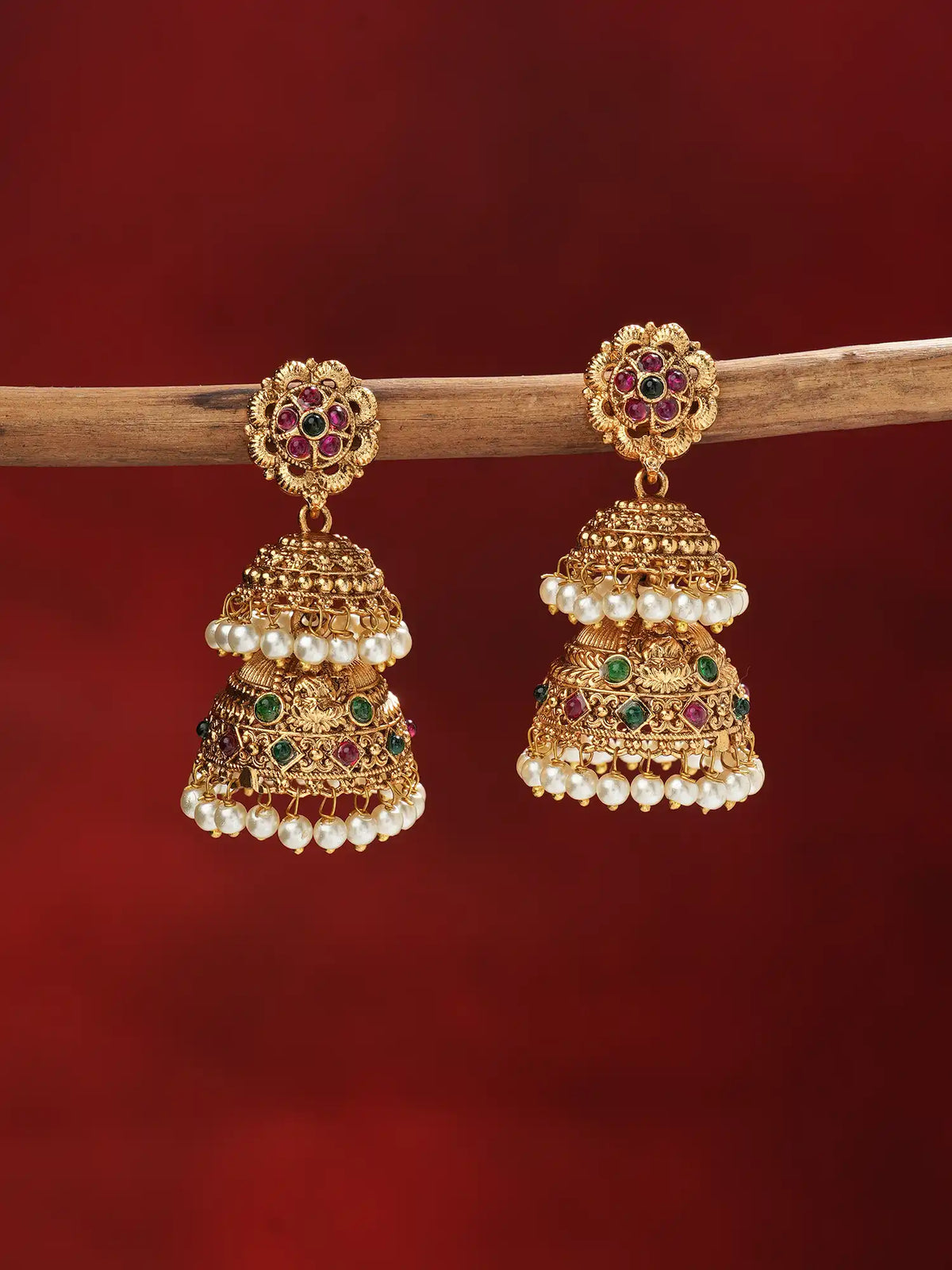 A pair of ornate gold-toned jhumka earrings with red and green stone accents and rows of white pearl beads, hanging from a wooden branch against a deep red background.
