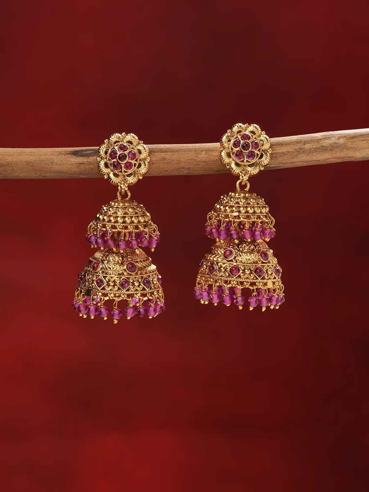 A pair of traditional Indian gold-plated jhumka earrings with pink gemstone accents, hanging from a wooden branch against a dark red background.