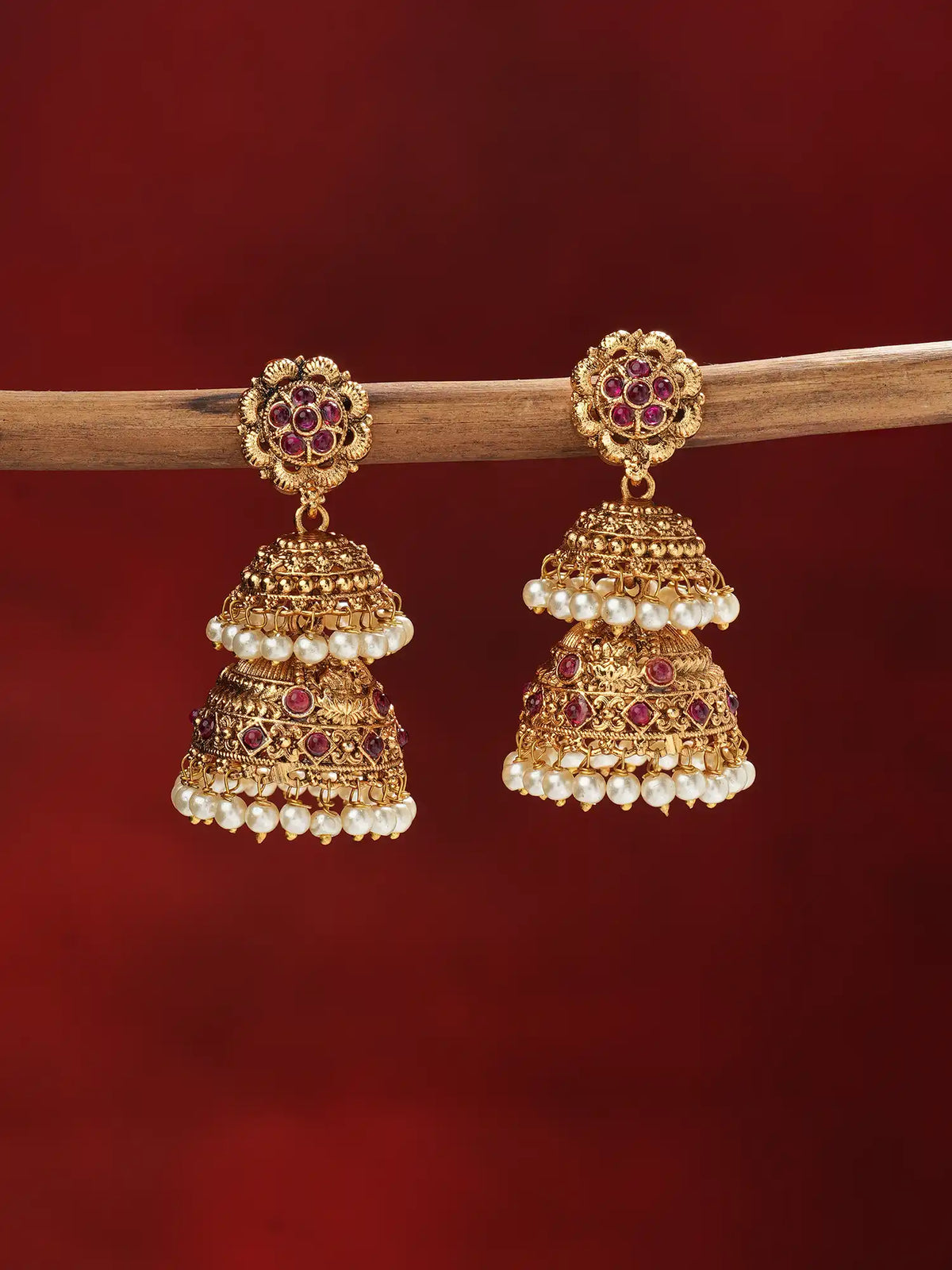A pair of ornate gold-toned jhumka earrings with red gemstone accents and white pearl bead borders, hanging from a wooden branch against a deep red background.