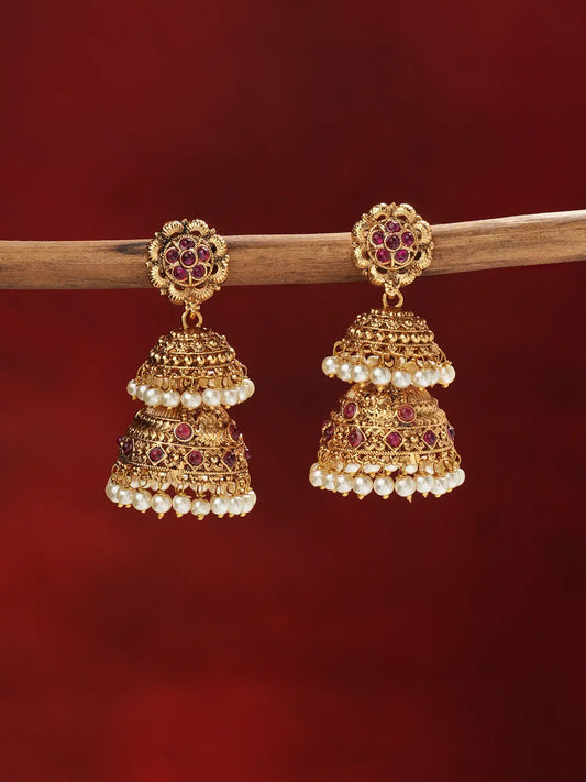 A pair of ornate gold-toned jhumka earrings with red gemstone accents and white pearl bead borders, hanging from a wooden branch against a deep red background.