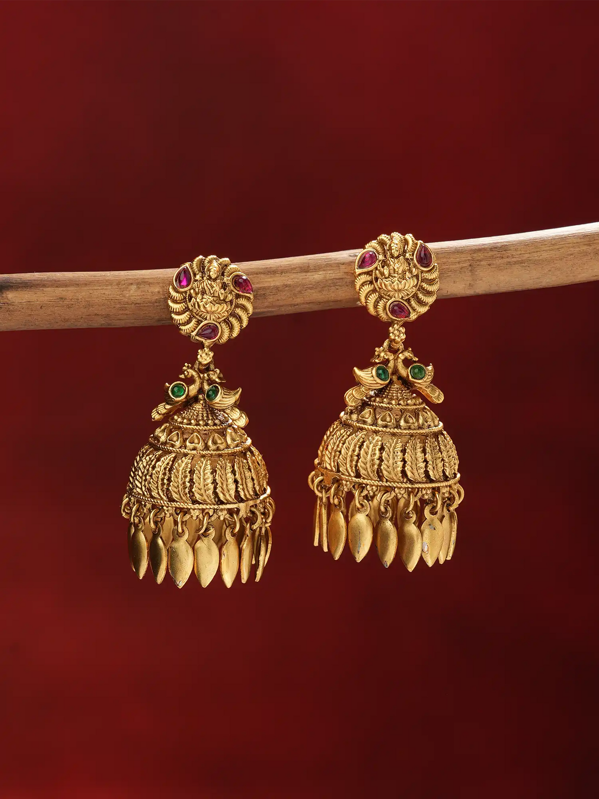 A pair of ornate gold-toned Indian jhumka earrings with small red and green stone accents, hanging from a wooden branch against a deep red background.