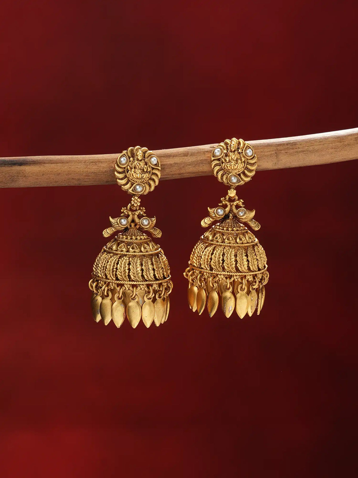 A pair of ornate gold-toned traditional jhumka earrings with small pearl-like accents, hanging from a wooden branch against a deep red background.