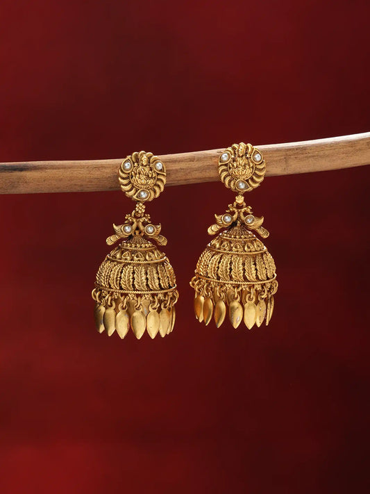 A pair of ornate gold-toned traditional jhumka earrings with small pearl-like accents, hanging from a wooden branch against a deep red background.