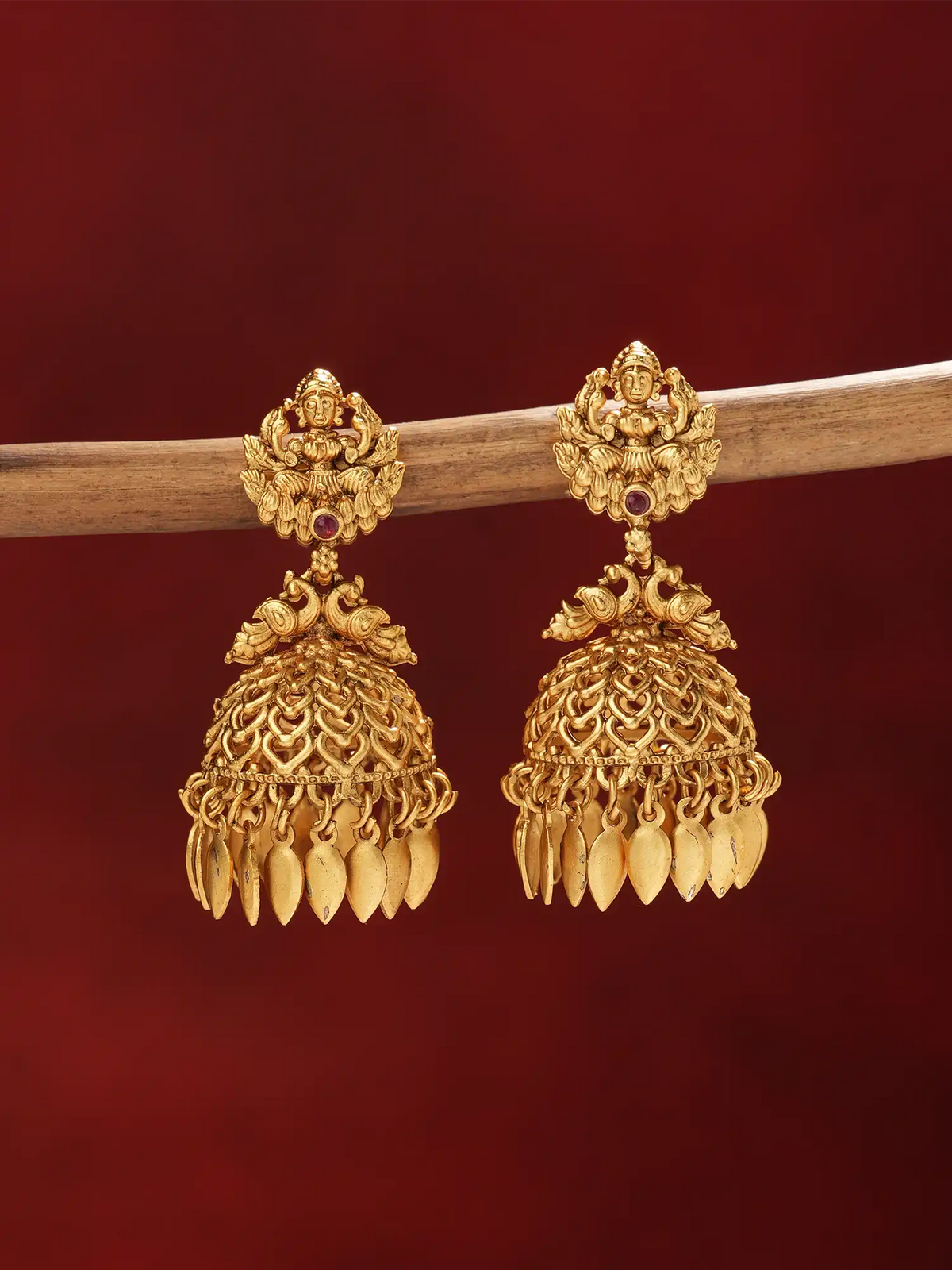 A pair of ornate gold-toned traditional jhumka earrings hanging from a horizontal wooden branch against a deep red background.