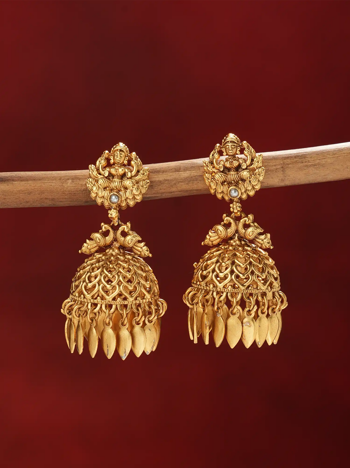 A pair of ornate gold-toned jhumka earrings hanging from a wooden branch against a deep red background.