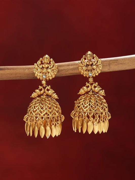 A pair of ornate gold-toned jhumka earrings hanging from a wooden branch against a deep red background.