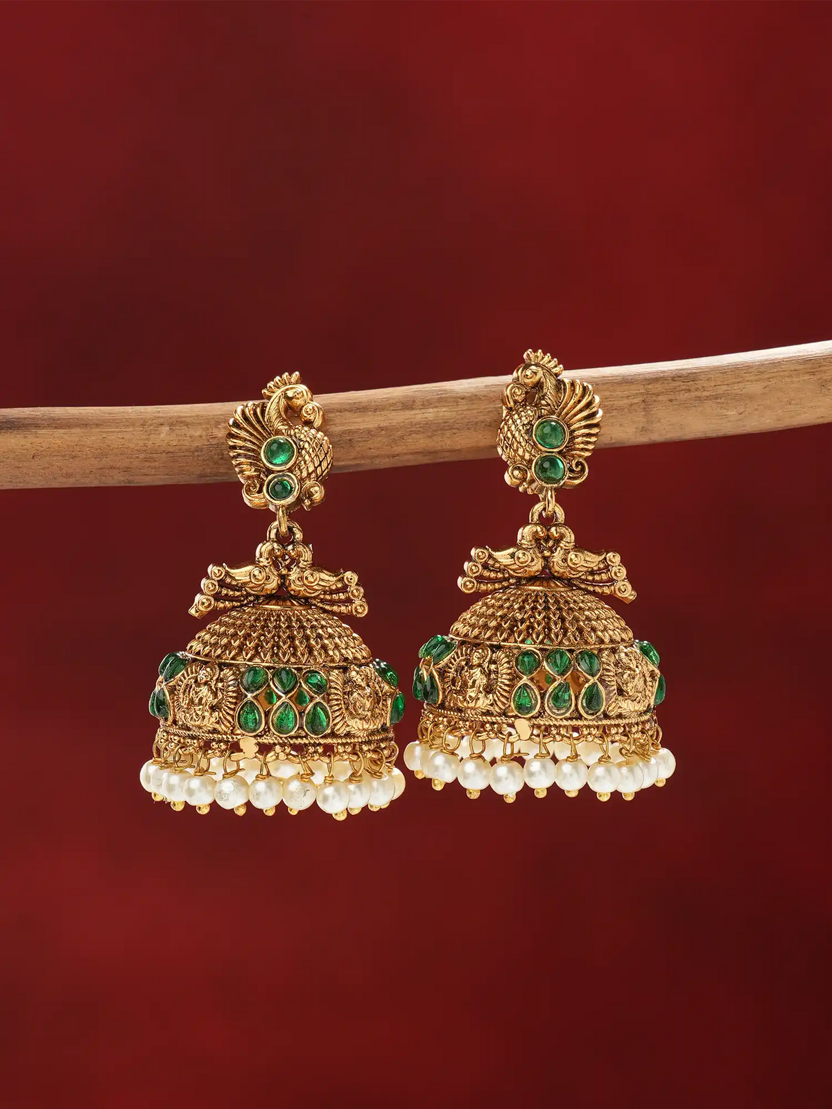 A pair of ornate gold-toned jhumka earrings with green stone accents and dangling white pearl fringe, hanging from a wooden branch against a deep red background.