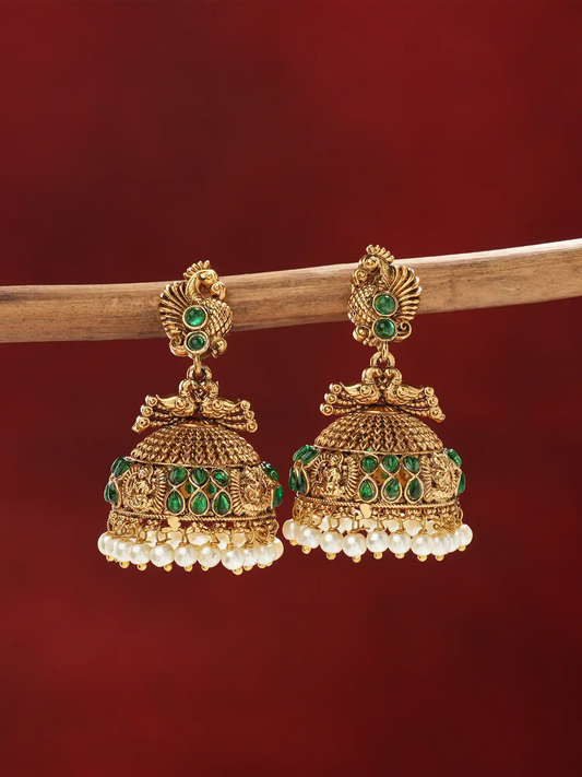 A pair of ornate gold-toned jhumka earrings with green stone accents and dangling white pearl fringe, hanging from a wooden branch against a deep red background.