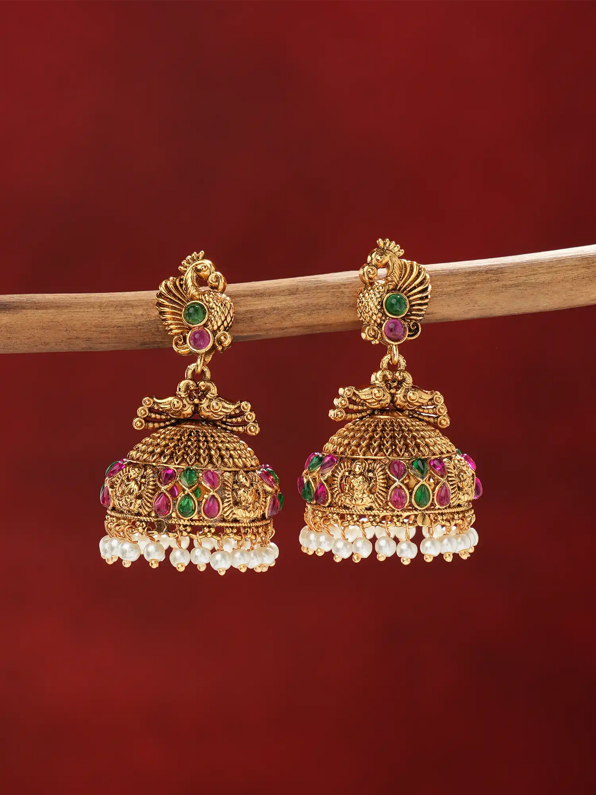 A pair of ornate gold-toned jhumka earrings with green and pink stone accents and dangling pearl-like beads, hanging from a wooden branch against a deep red background.
