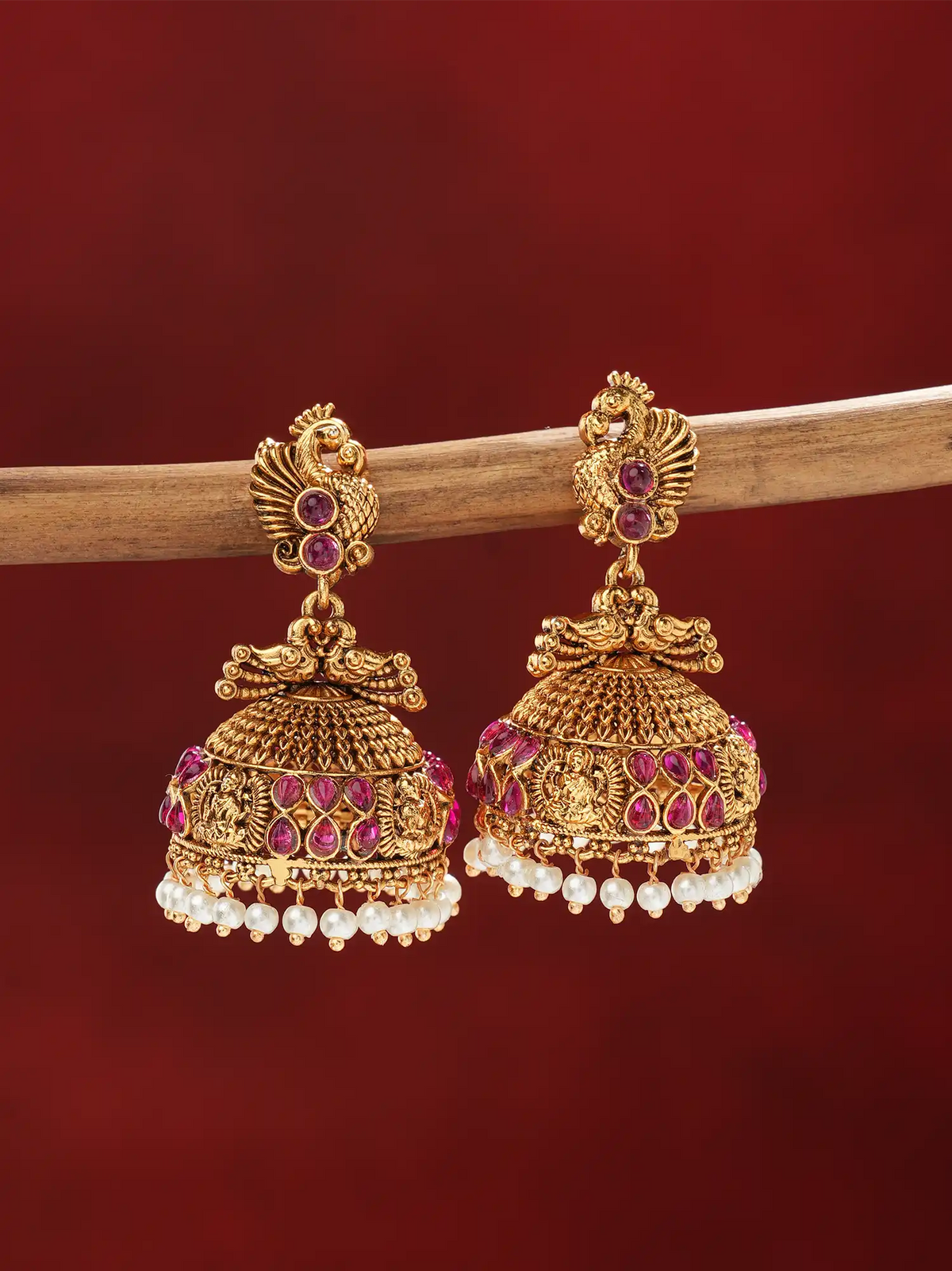 A pair of ornate gold-toned jhumka earrings with pink gemstone accents and dangling white pearl beads, displayed on a wooden branch against a deep red background.