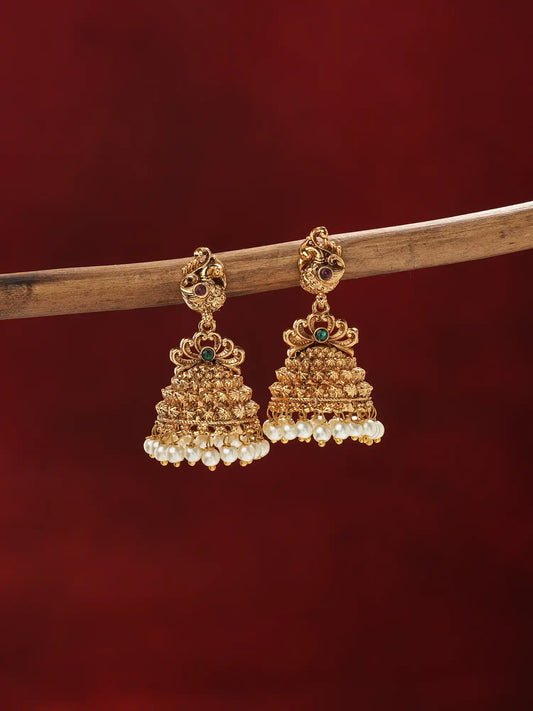 A pair of ornate gold-toned jhumka earrings with small green stone accents and rows of white pearl-like beads at the bottom, hanging from a wooden branch against a dark red background.