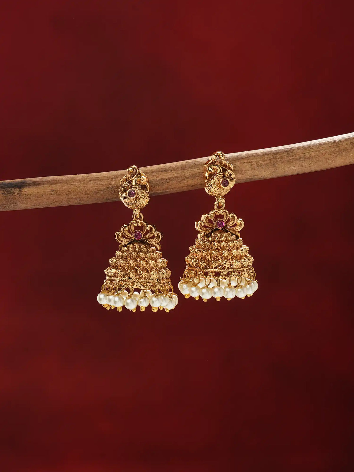 A pair of ornate gold-toned jhumka earrings with small red gemstone accents and rows of white pearl-like beads at the bottom, hanging from a wooden branch against a dark red background.