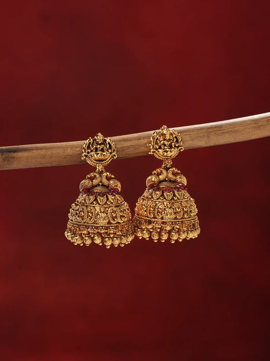 A pair of ornate gold-toned Indian jhumka earrings hanging from a wooden branch against a dark red background.