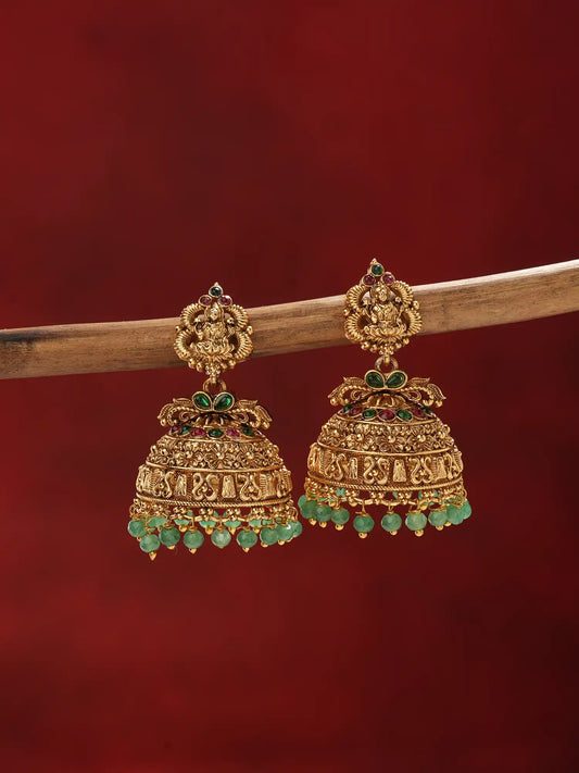 A pair of ornate gold-toned jhumka earrings with green stone accents and dangling green beads, displayed against a dark red background with a wooden branch behind them.