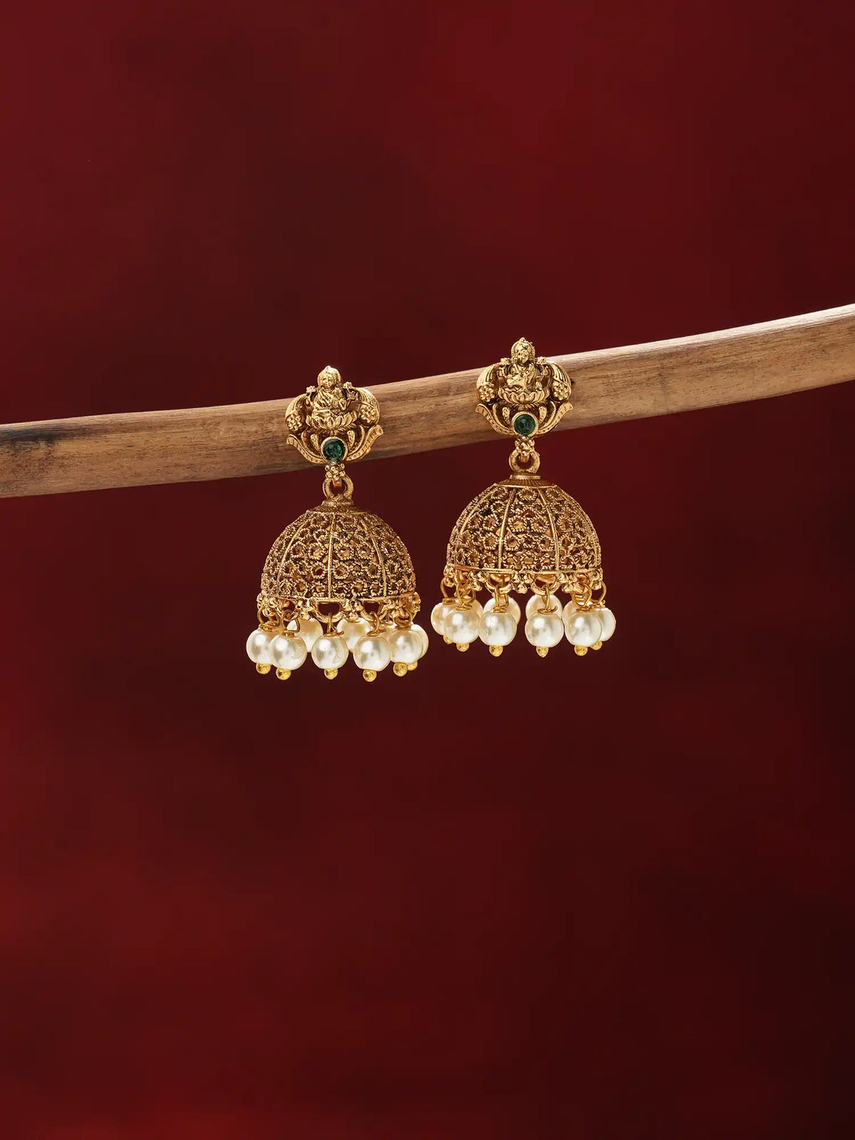 A pair of traditional Indian gold-plated jhumka earrings with white pearl bead drops and small green stone accents, hanging from a wooden branch against a dark red background.