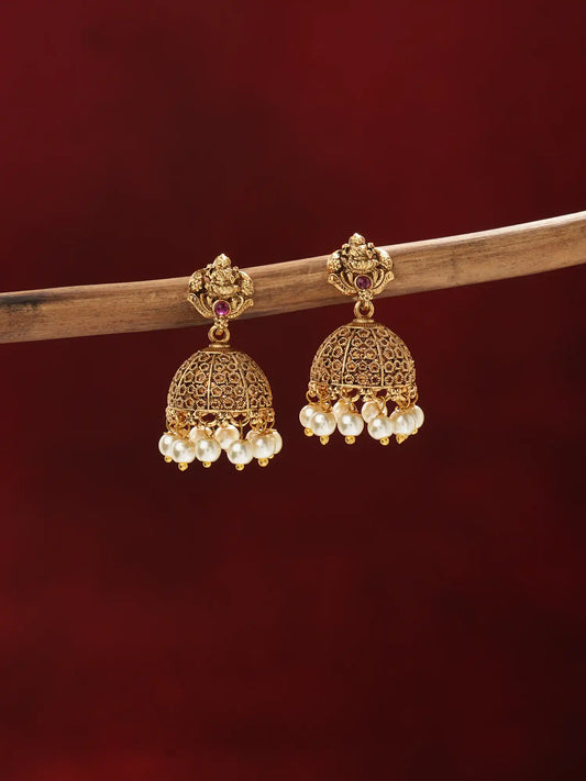 A pair of ornate gold-toned jhumka earrings with small red stones and clusters of white pearl-like beads, hanging from a wooden branch against a dark red background.
