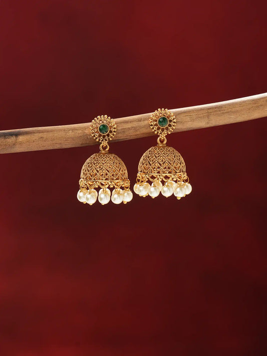 A pair of gold-toned traditional Indian jhumka earrings with green stone accents and dangling pearl beads, hanging from a wooden branch against a dark red background.