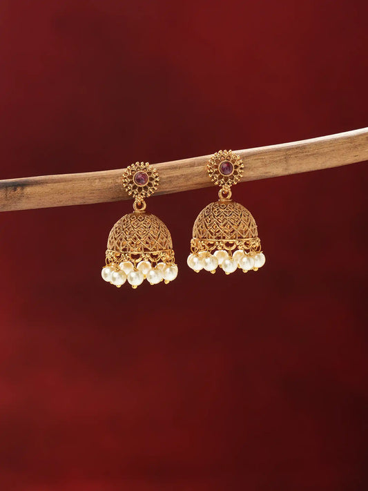 A pair of ornate gold-toned jhumka earrings with small purple stones at the top and rows of white pearl-like beads hanging from the bottom, displayed on a wooden branch against a dark red background.