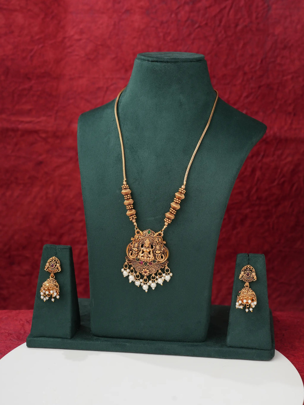 Gold-toned traditional Indian jewelry set featuring a necklace and matching earrings displayed on a dark green velvet jewelry bust against a textured red background.