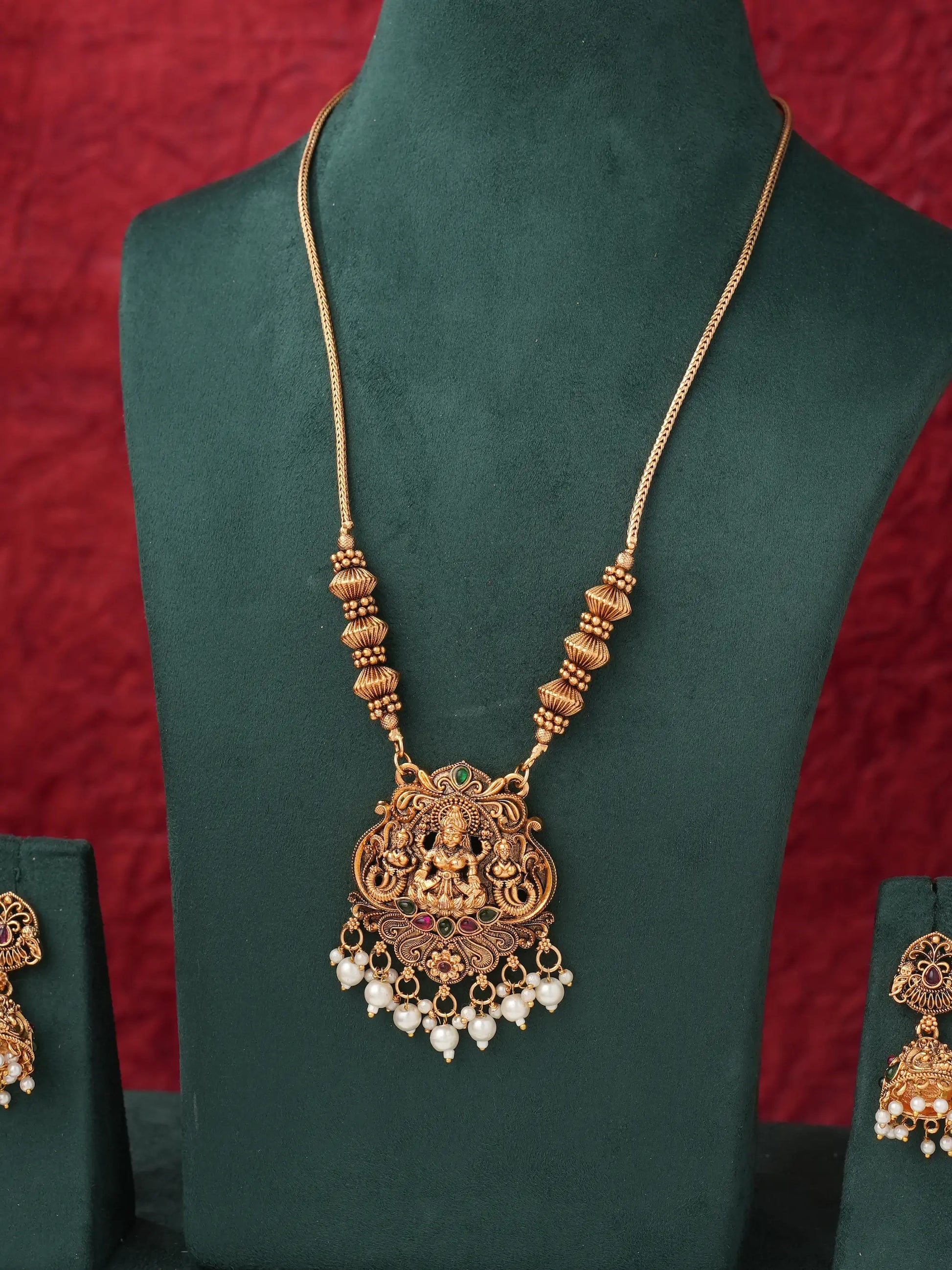 An ornate gold-toned necklace with a detailed Ganesha pendant, displayed on a dark green velvet jewelry bust against a textured red background. The necklace features a chain, decorative beads, and dangling white pearl-like accents.