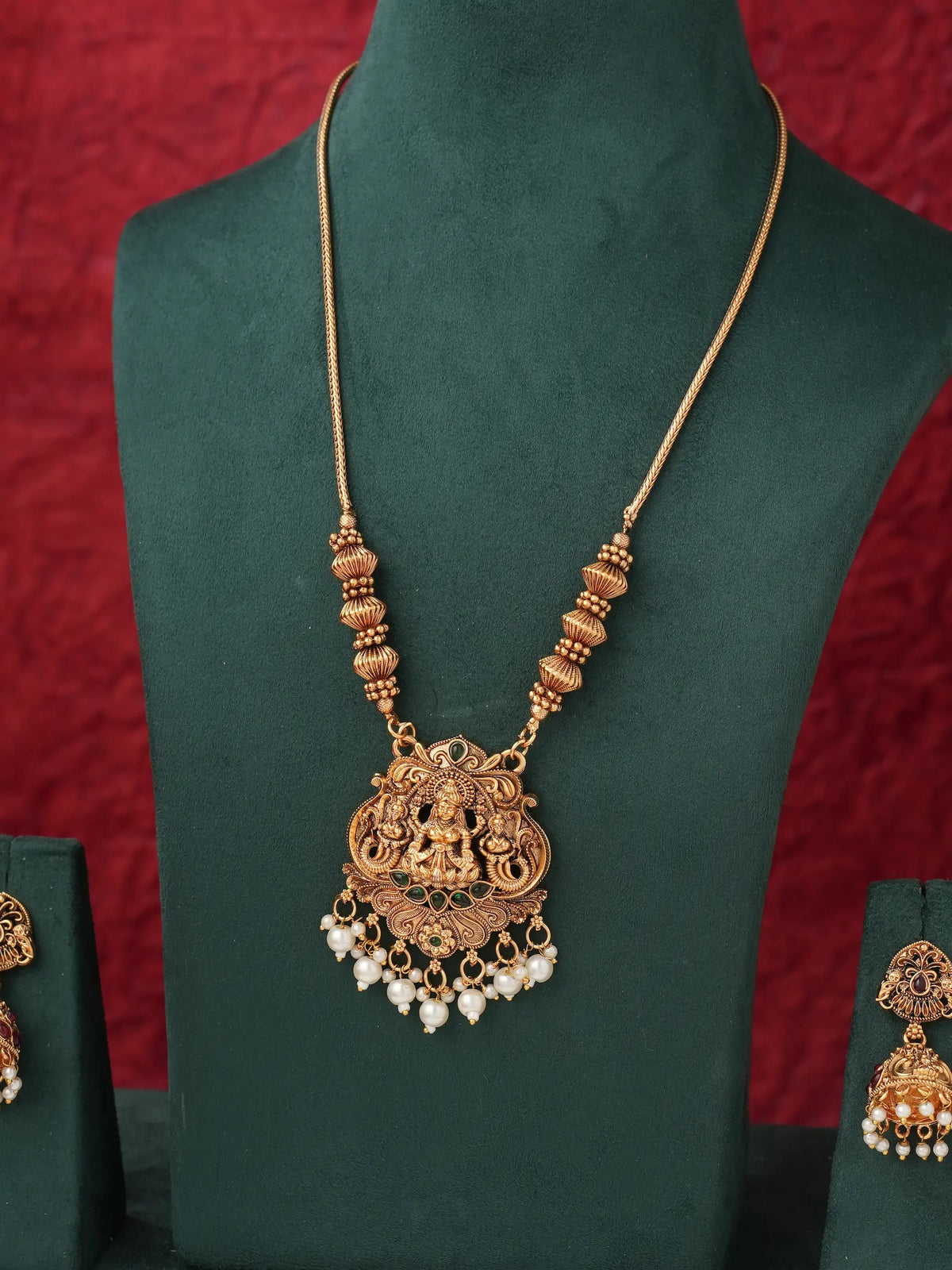 An ornate gold-toned necklace with a detailed central pendant and dangling pearl-like beads, displayed on a dark green jewelry bust against a textured red background.