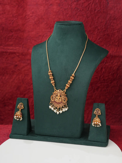 An ornate gold-toned traditional necklace with a detailed pendant and matching drop earrings displayed on a dark green jewelry bust set against a textured red backdrop.
