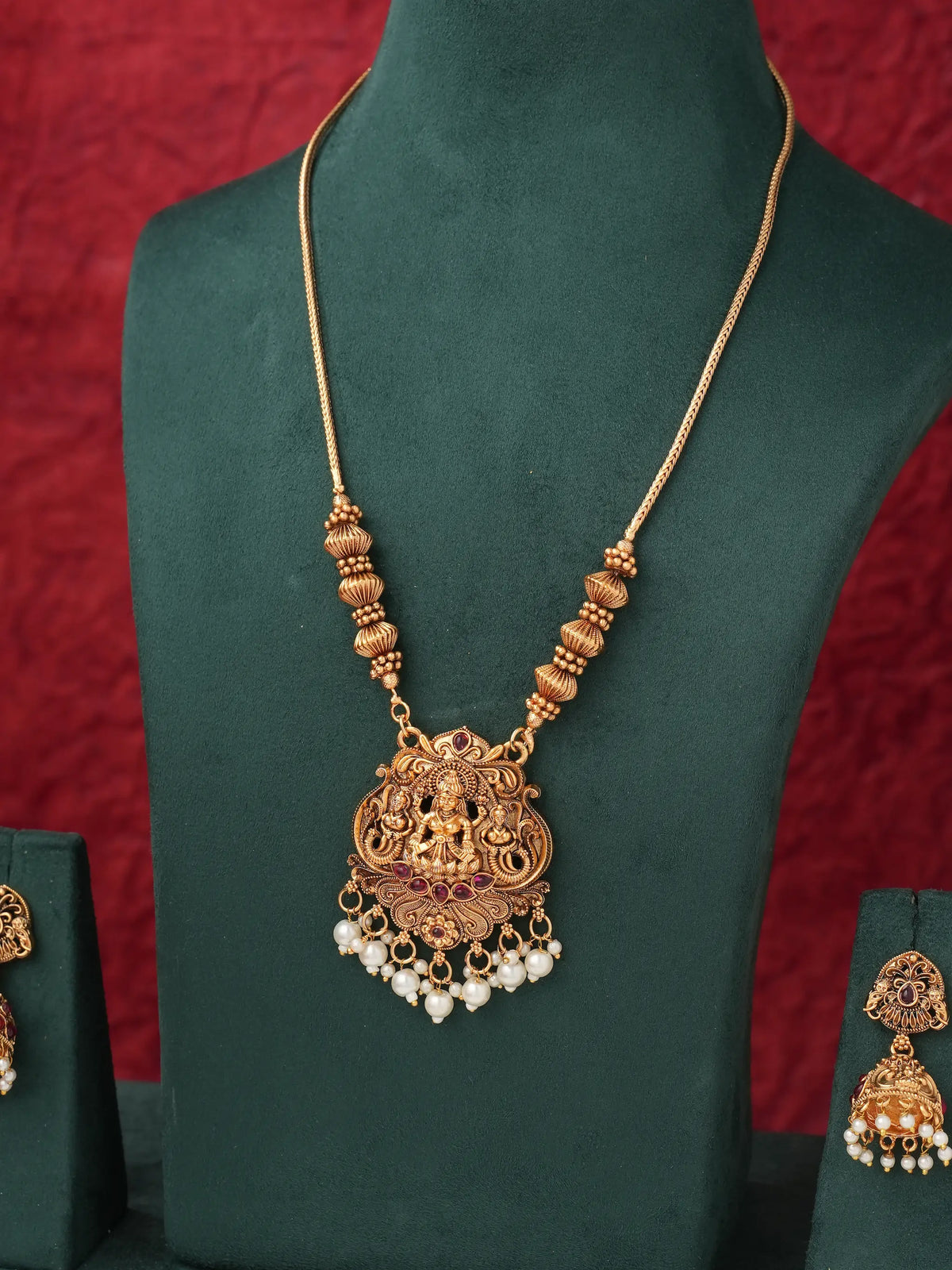 An ornate gold-toned necklace with a detailed pendant featuring a deity motif, red stone accents, and dangling pearl-like beads, displayed on a dark green jewelry bust against a textured red background.