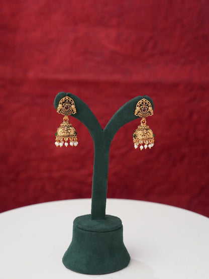 A pair of ornate gold-toned Indian jhumka earrings displayed on a dark green velvet jewelry stand against a textured red background.