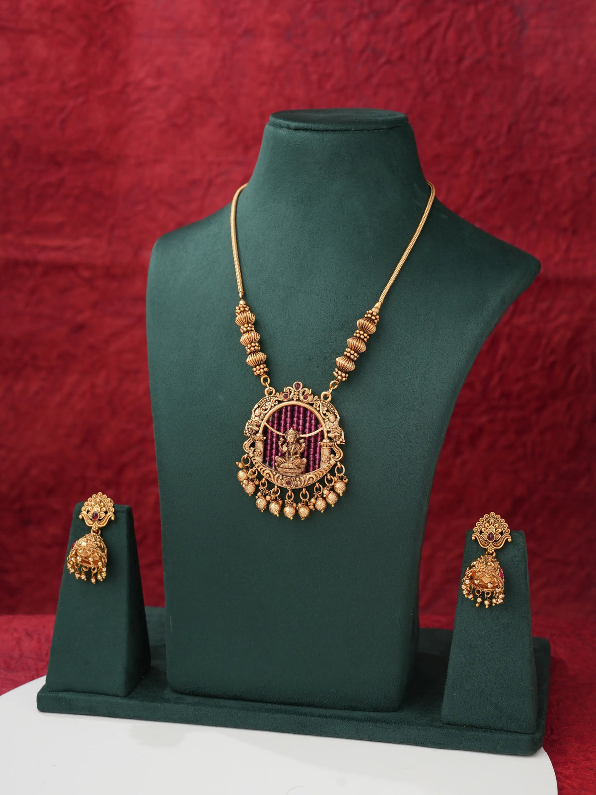 Gold-toned jewelry set displayed on a dark green mannequin bust, featuring a necklace with a circular pendant and matching earrings, set against a textured red backdrop.