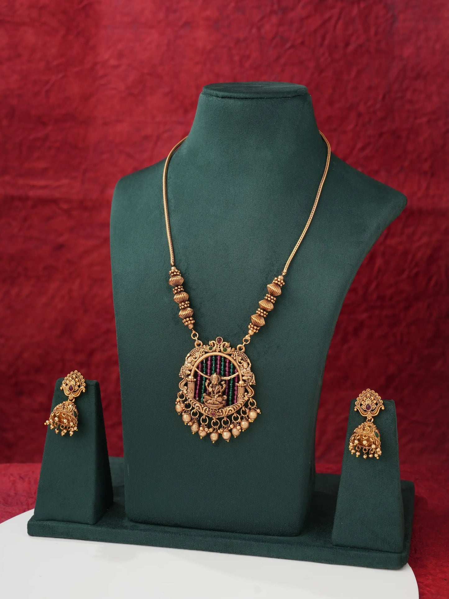 Gold-toned traditional Indian jewelry set featuring a necklace and matching earrings displayed on a dark green velvet jewelry stand against a textured red backdrop.