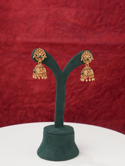 A pair of gold-toned traditional Indian jhumka earrings with green stone accents, displayed on a dark green velvet jewelry stand against a textured red background.