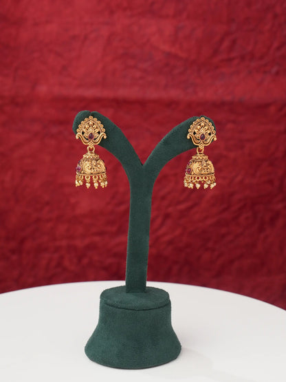 A pair of ornate gold-toned jhumka earrings displayed on a dark green Y-shaped jewelry stand against a textured red background.