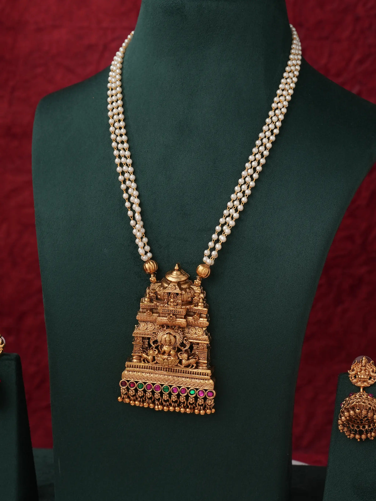 An ornate gold-toned necklace with a temple-inspired pendant and a dual-strand pearl chain, displayed on a dark green jewelry bust against a red textured background.