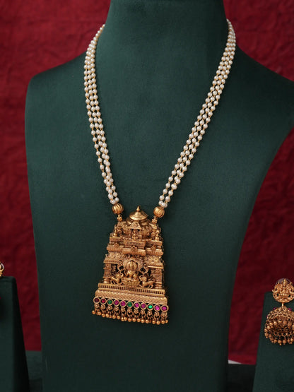 An ornate gold-toned necklace with a temple-inspired pendant and a dual-strand pearl chain, displayed on a dark green jewelry bust against a red textured background.