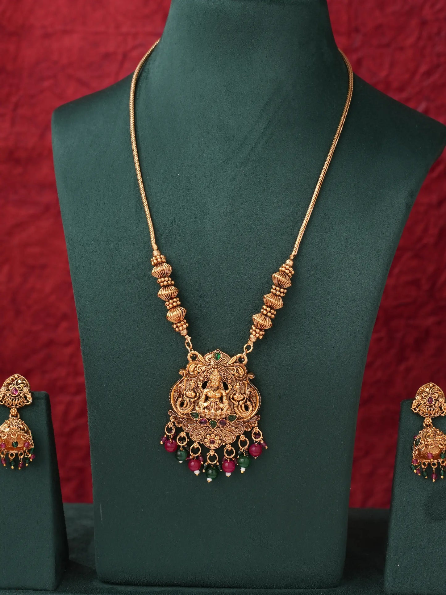 Gold-toned traditional necklace with a detailed pendant featuring a deity motif and red and green bead accents, displayed on a dark green jewelry bust with matching earrings partially visible on either side against a red textured backdrop.