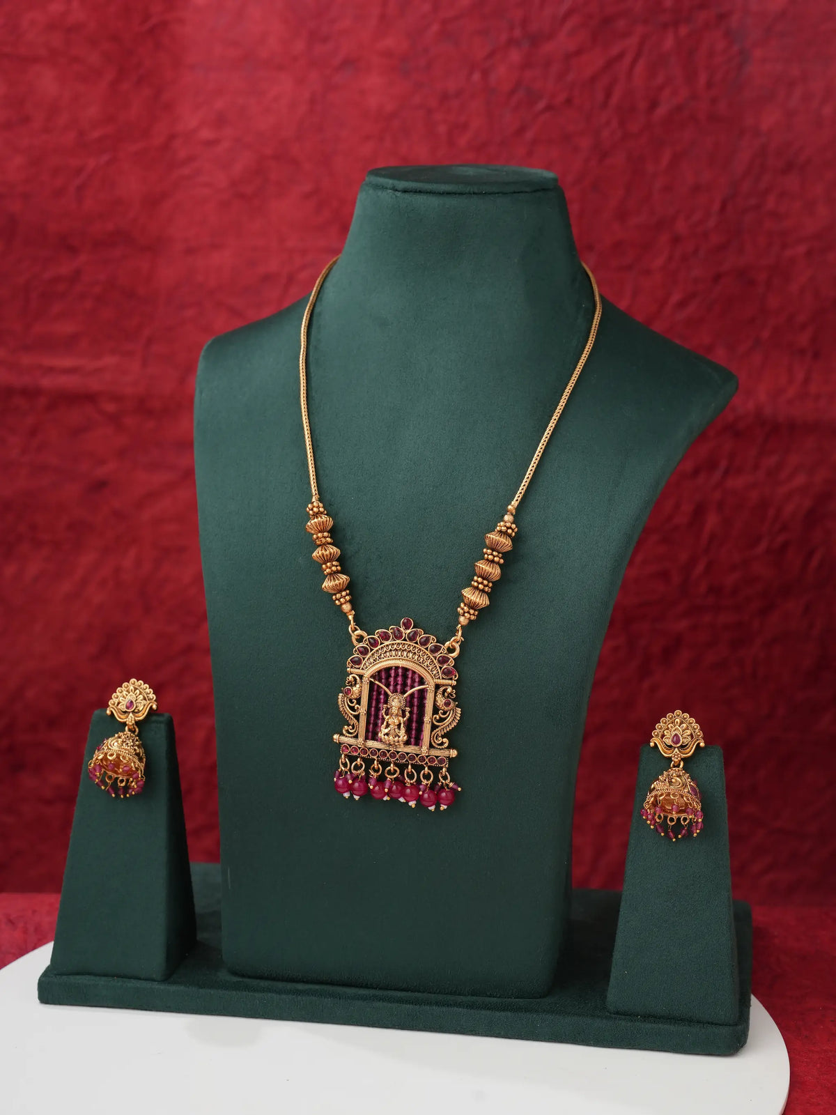 Gold-toned traditional Indian jewelry set featuring a necklace and matching earrings displayed on a dark green jewelry bust against a textured red backdrop.