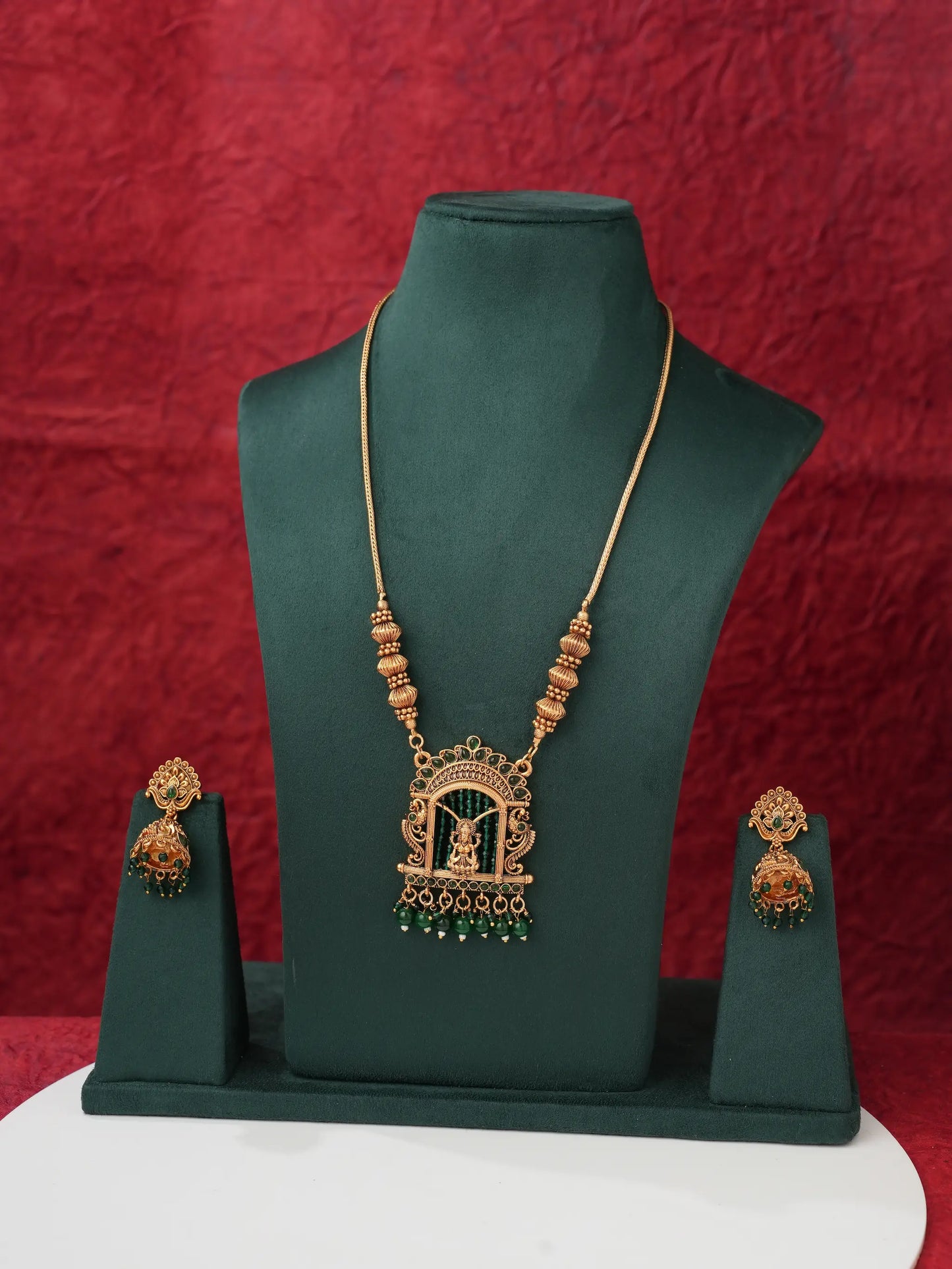Gold-toned traditional Indian jewelry set featuring a necklace with a temple arch pendant, matching earrings, and a matching necklace stand, displayed on a dark green mannequin bust against a textured red background.