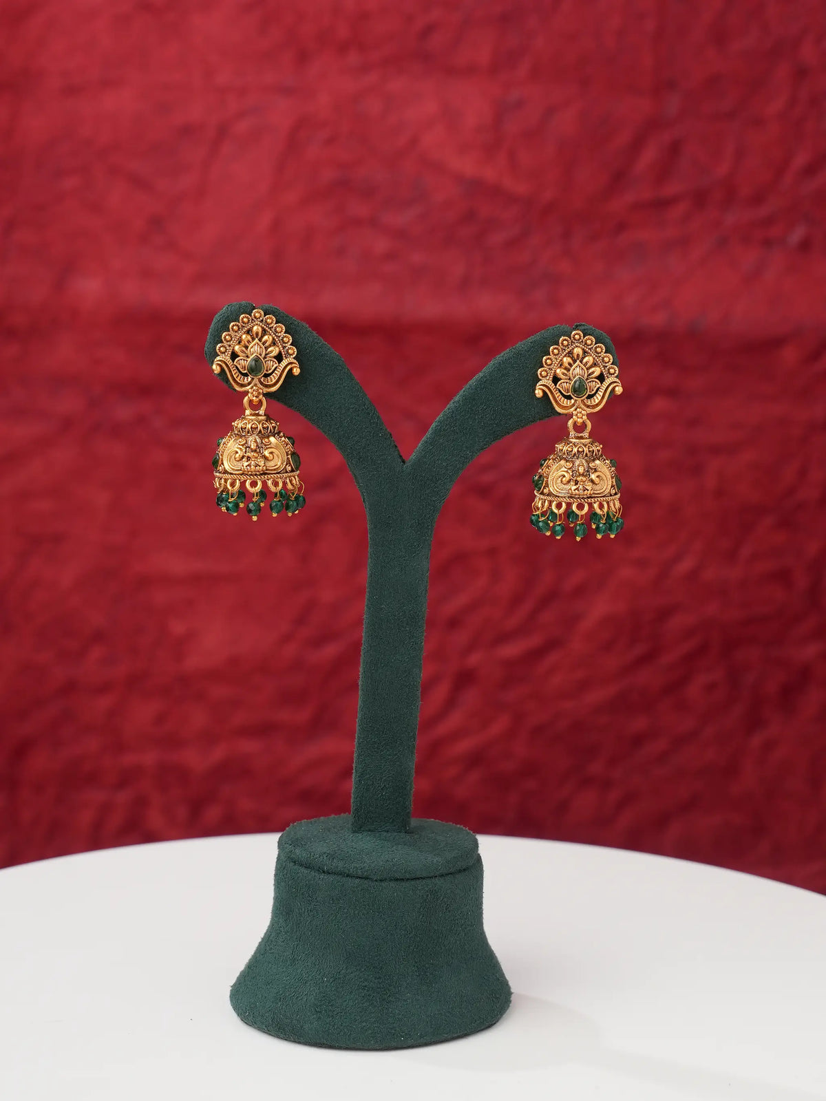 A pair of traditional Indian gold jhumka earrings with green stone accents, displayed on a dark green velvet jewelry stand against a textured red background.