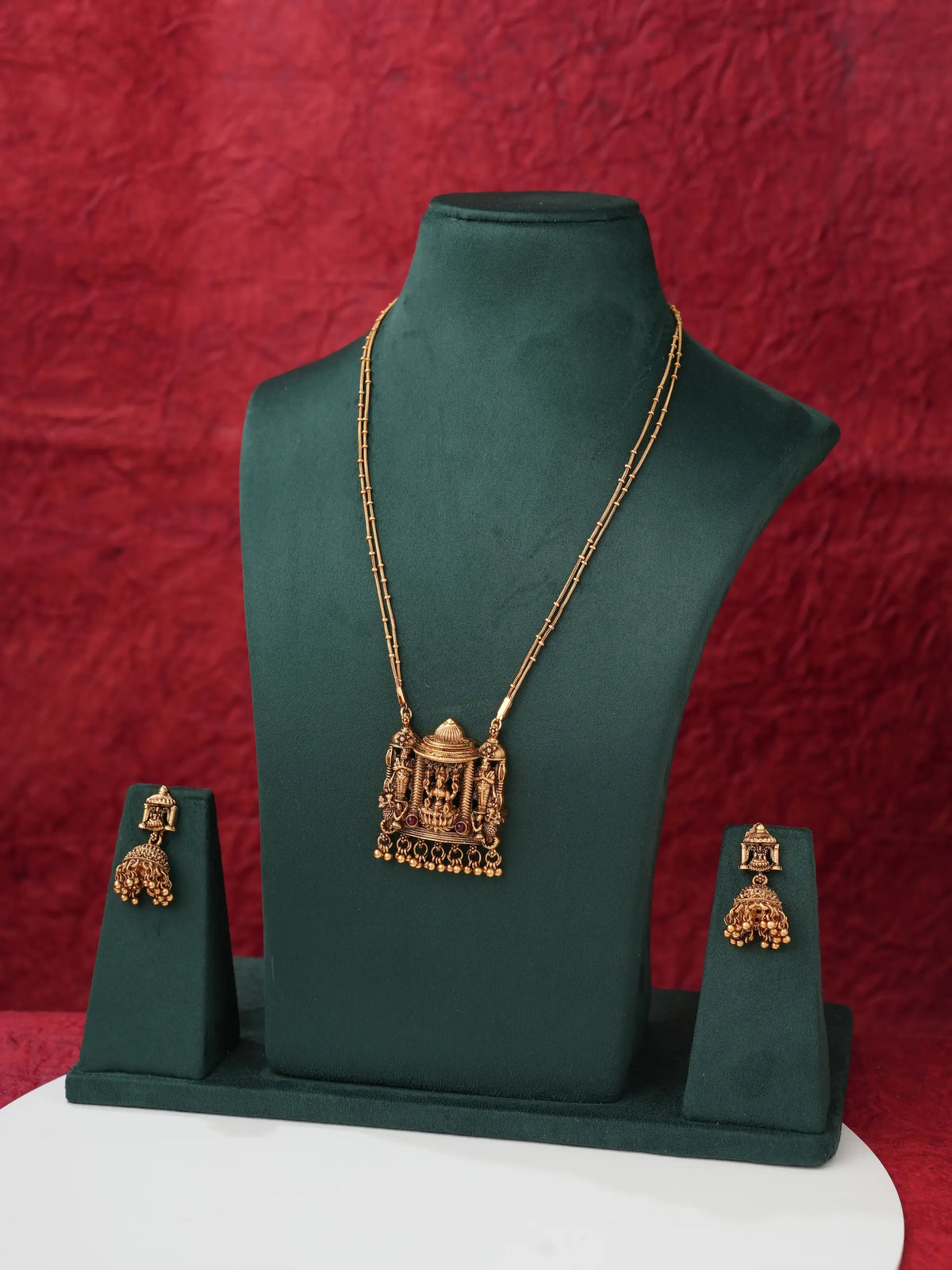 Gold-toned jewelry set displayed on a dark green mannequin bust, including a necklace with an ornate pendant and matching earrings, set against a textured red backdrop.