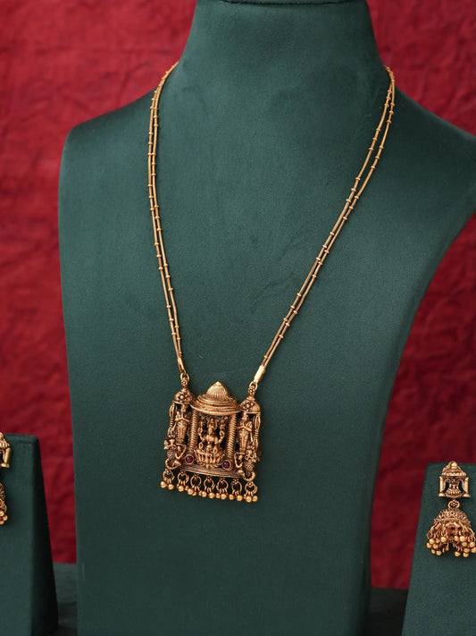 An ornate gold-toned necklace with a detailed pendant displayed on a dark green jewelry bust, with matching earrings partially visible on side stands against a textured red background.