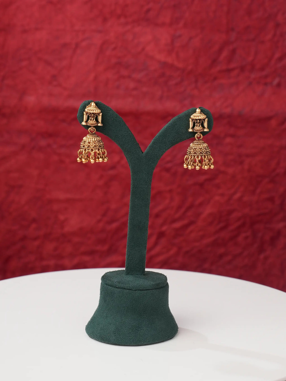 A pair of gold-toned traditional Indian jhumka earrings displayed on a dark green velvet jewelry stand against a textured red background.