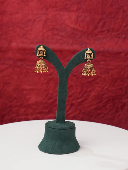 A pair of gold-toned traditional Indian jhumka earrings displayed on a dark green velvet jewelry stand against a textured red background.