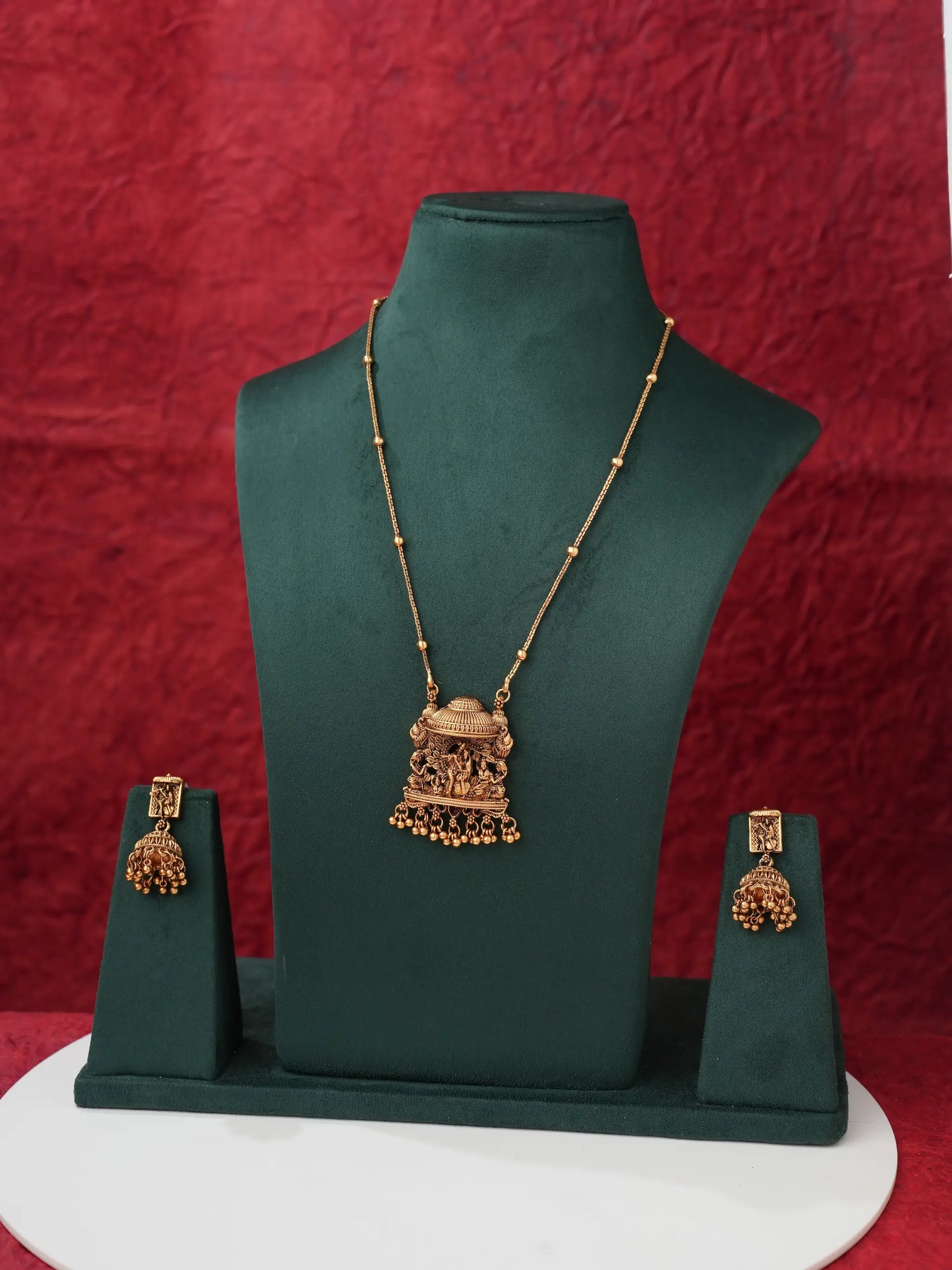 Gold-toned traditional Indian jewelry set featuring a necklace with a detailed pendant and matching earrings, displayed on a dark green jewelry bust against a textured red background.